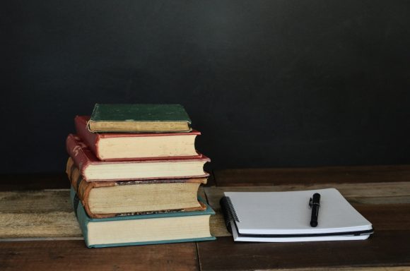 Stack of old books next to a blank notepad and a pen on a wooden surface.